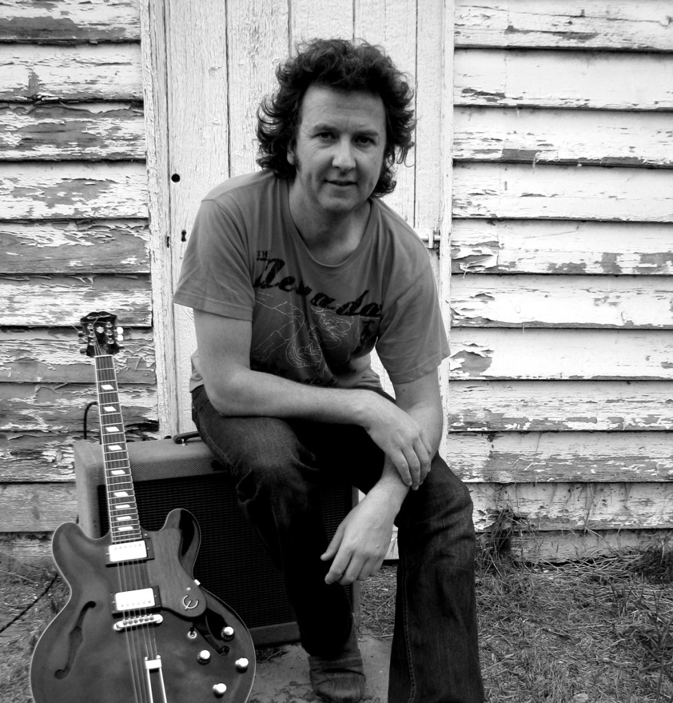 Patrick Shawcross seated with his hollow-body guitar against a weatherboard wall